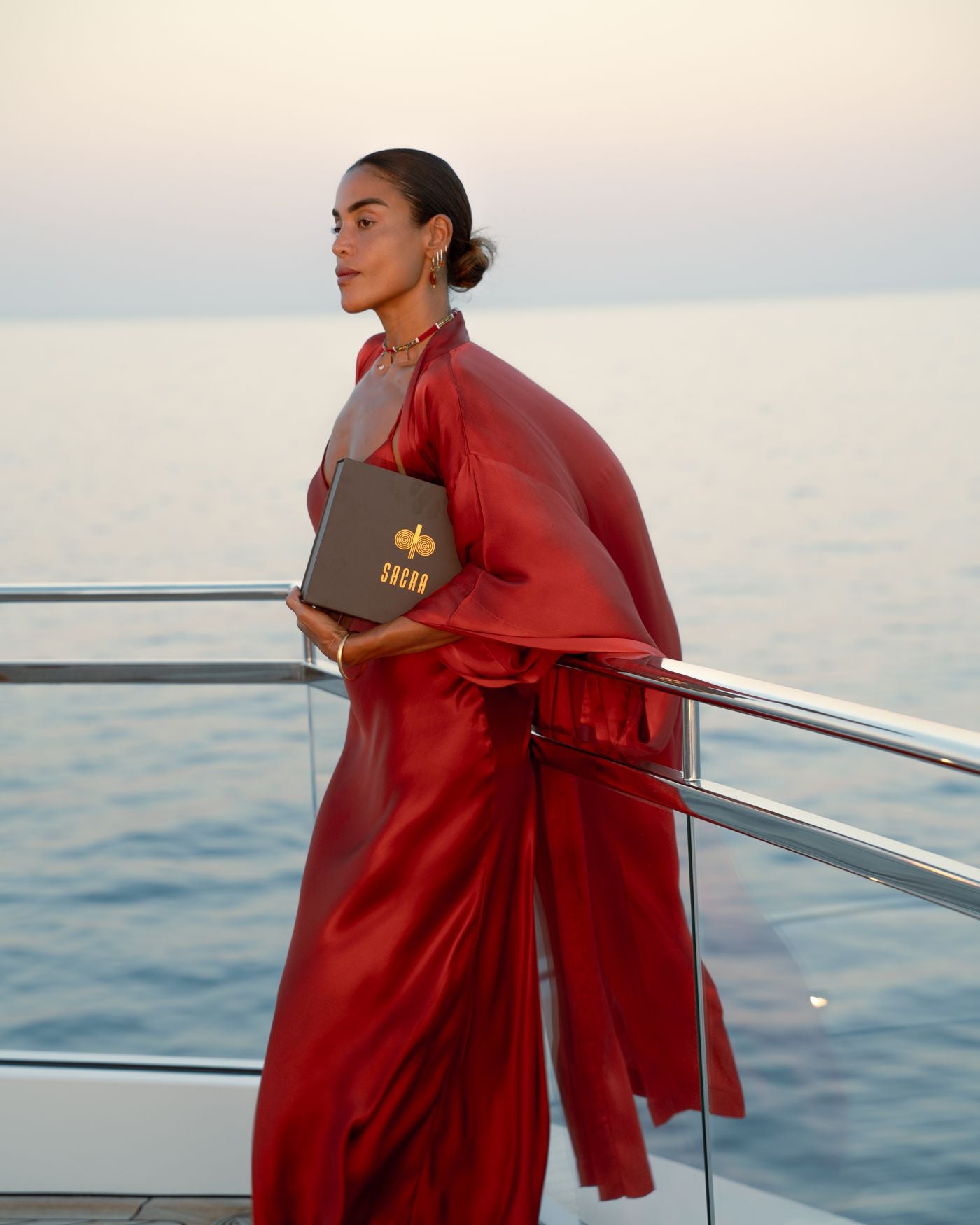 A woman in red silk at a boat rail at sunset, holding the SACRA box